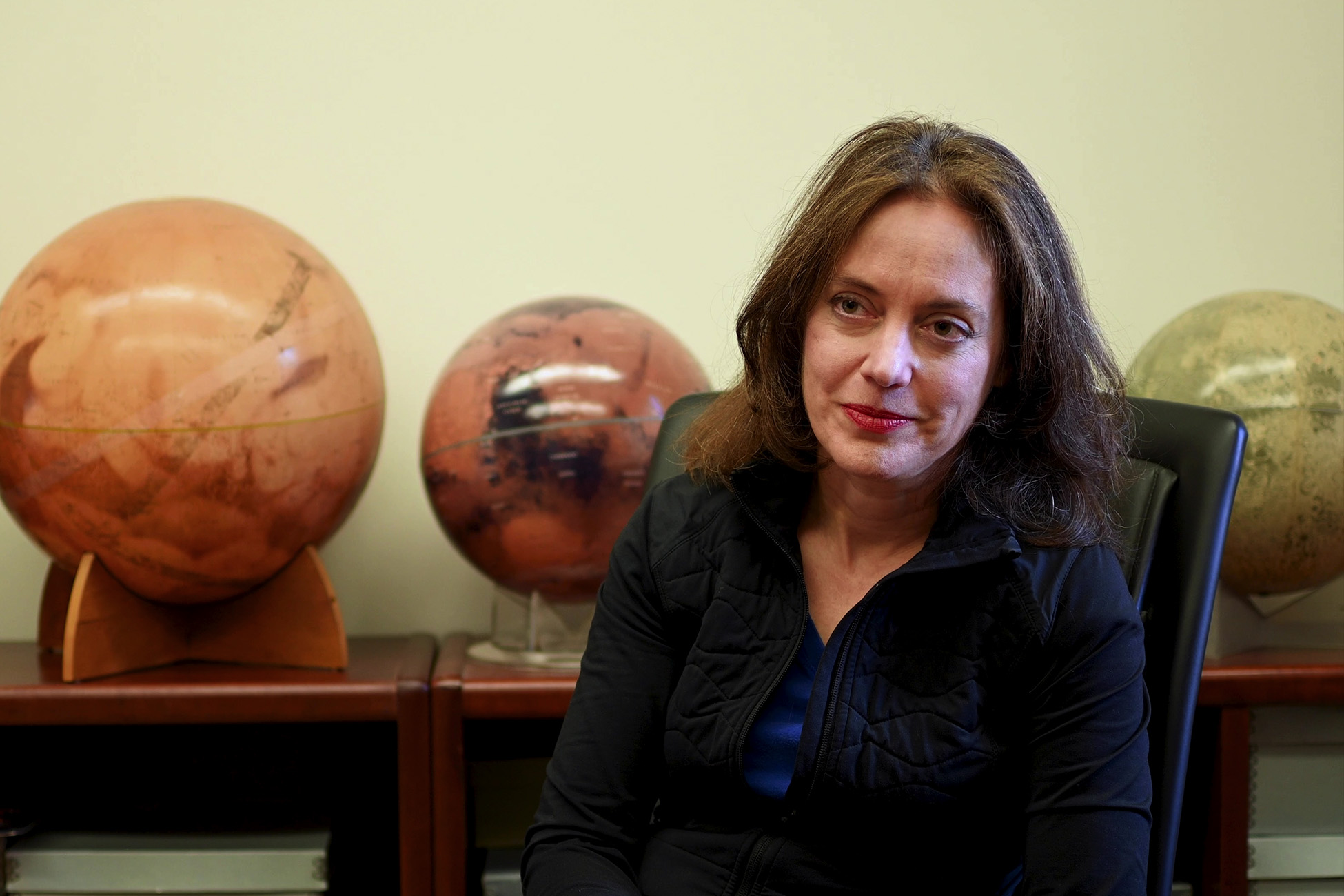 A woman sits in an office chair, looking slightly to the side, with three spherical Mars globes displayed on shelves behind her.